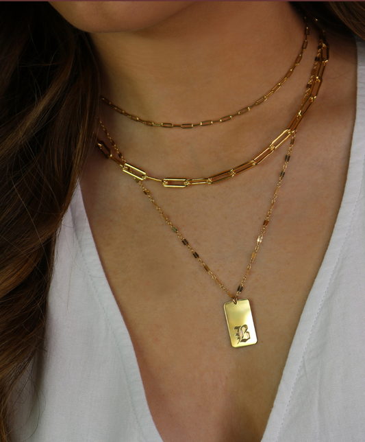 Gold necklaces with a tag pendant on a person wearing a white top, branded 'Gems in Vogue'.